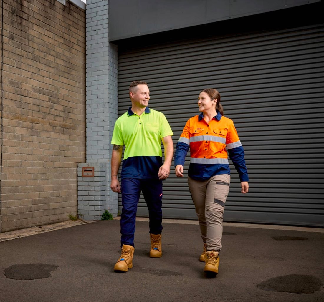 A smiling male and a female walk side by side in Hard Yakka Hi Vis, cargo pants and side zip safety boots