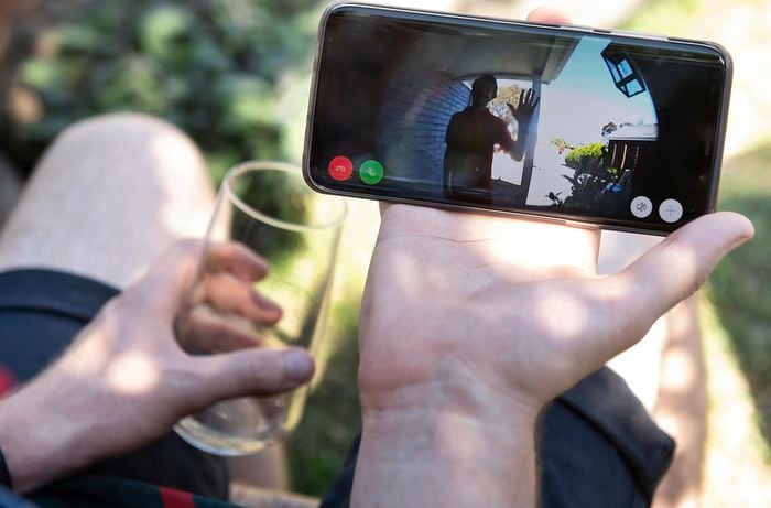 Person holding a glass and looking at their smart phone, which is feeding through vision from their door bell camera.