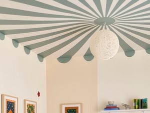 Ceiling mural painting with green and white radial stripes and a scalloped edge in a room corner, with a pendant light, cream walls, framed artwork and a bookshelf.
