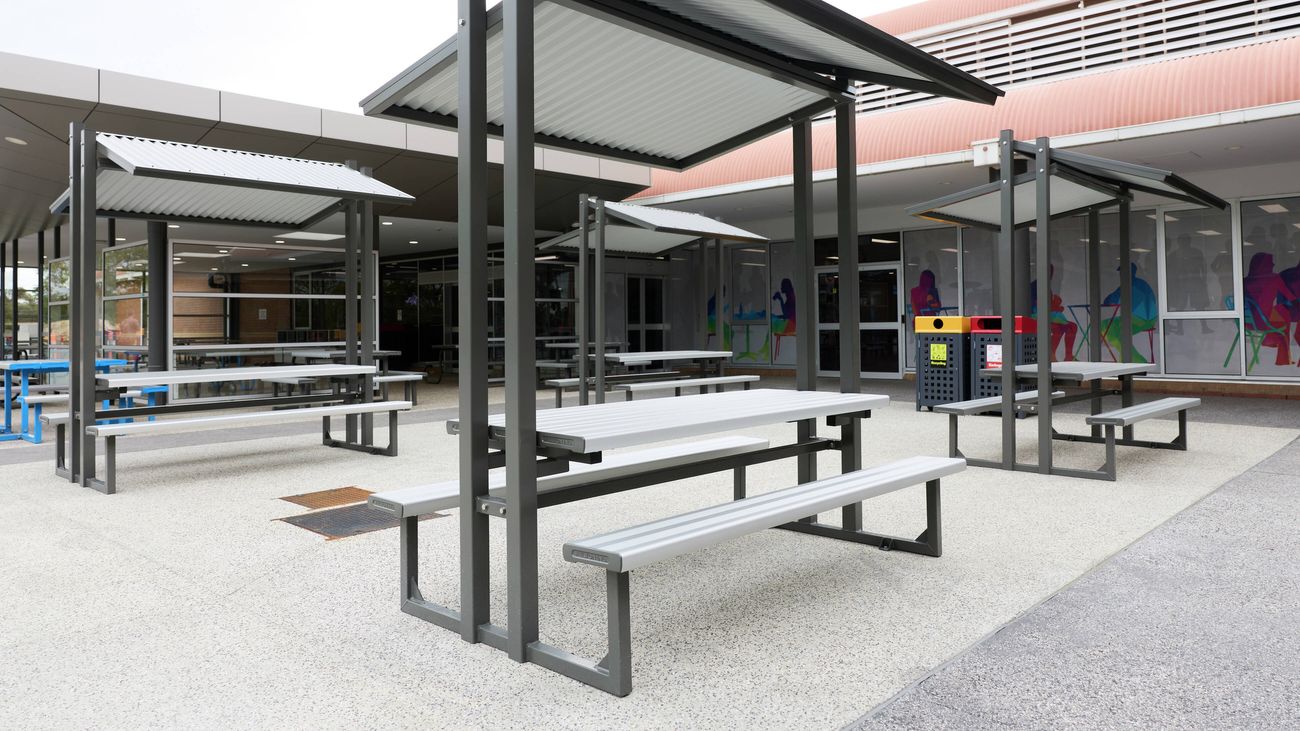 Outdoor covered seating area with steel-framed picnic tables and shelters in a school or campus courtyard setting.