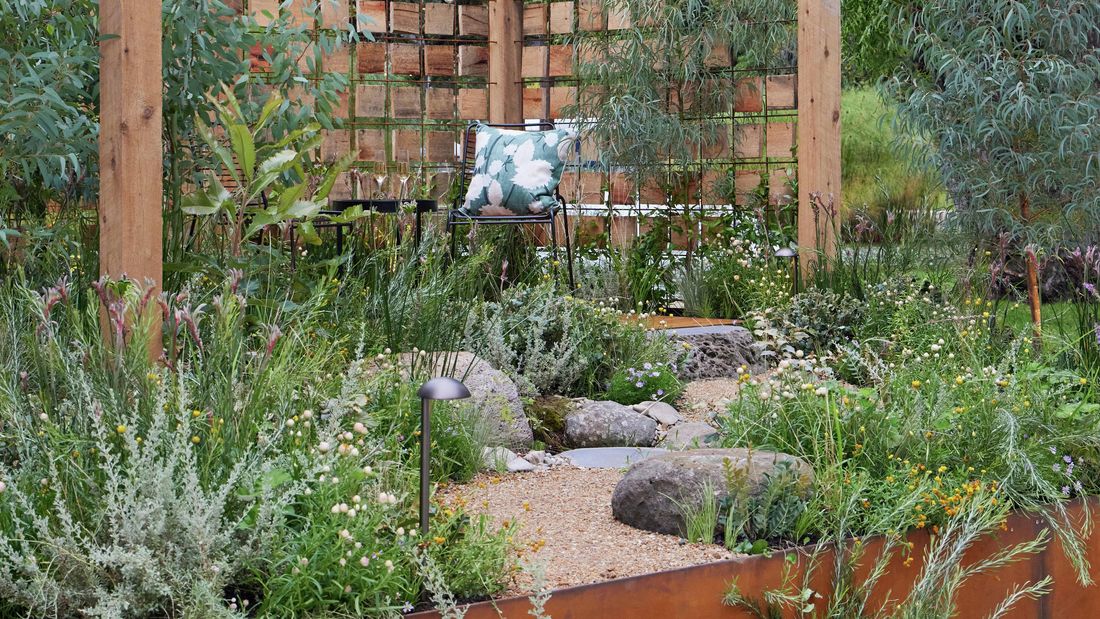 A garden corner with a timber and wire mesh trellis, surrounded by lush native plants, rocks, and a chair with a patterned cushion