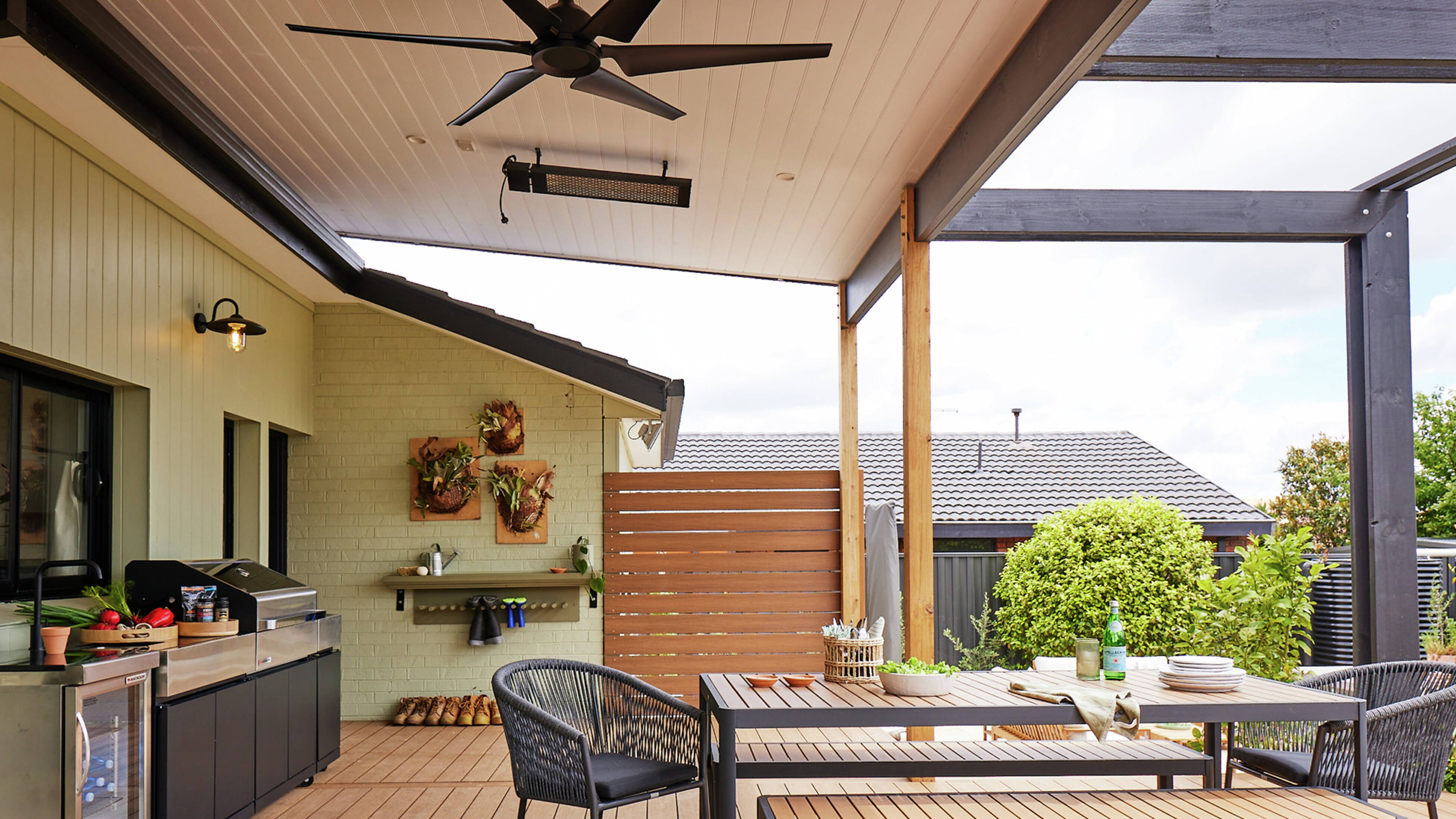  Outdoor dining setting with a table, benches and chairs beneath a dark timber pergola on a deck, with a garden view.]