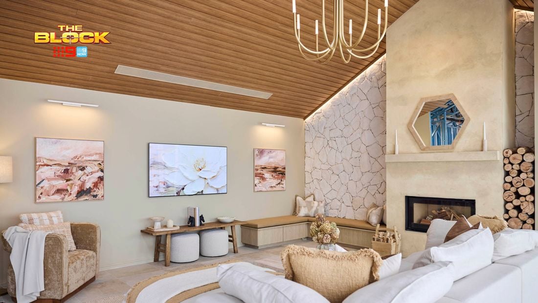 A modern living room with a high wooden ceiling and a large chandelier. The space includes a white and beige sectional sofa with pillows, an armchair draped with a blanket, and two small round stools. A fireplace on the right is framed by stacked firewood, candles, and an octagonal mirror above. The back wall features two paintings flanking a central TV displaying flowers. The left wall is clad in textured stone.