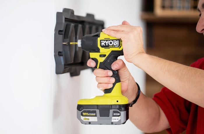 : A person is using a Ryobi cordless drill to secure a Ryobi LINK rail onto a garage wall with screws.  