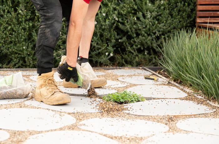 A person wearing work boots and gloves is spreading small pebbles into the gaps between stone pavers