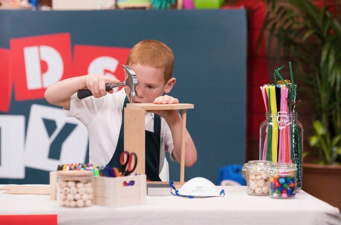 One kid is using a hammer to nail the base to the sides of a tool caddy in a Bunnings DIY workshop, with many arts and crafts supplies on the table.