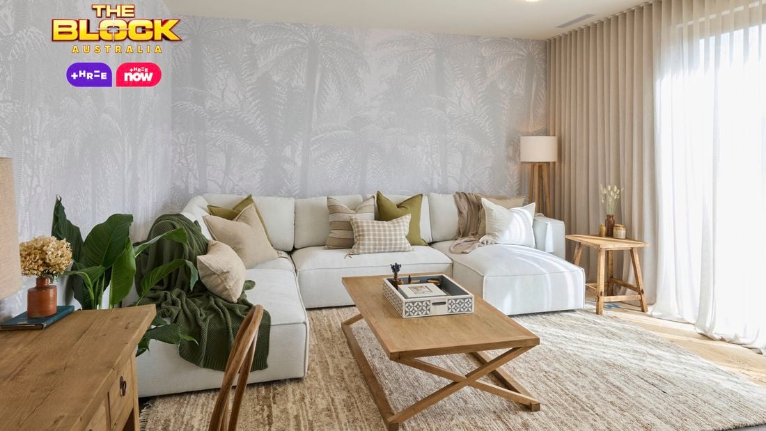 A modern living room with a large white sectional sofa decorated with neutral and green pillows. A wooden coffee table with a decorative tray sits in front, and a side table with a lamp and vase is placed to the right. Light-colored curtains cover large windows, and palm tree wallpaper adds texture to the walls. A desk with drawers and a chair is positioned on the left, near potted plants. 