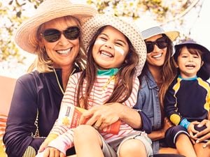 Two women both with young children, all wearing sun-smart clothing and hats, sitting outdoors. Adults wear sunglasses and children have zinc cream on their faces. One woman holds a tube of sunscreen.