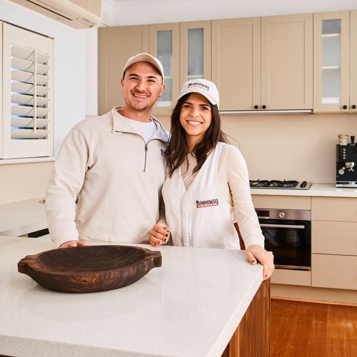   A couple consists of a man and a woman standing side by side behind a kitchen island in a modern beige kitchen, both wearing branded caps and casual tops, with cabinets, a cooktop, coffee machine, and stainless steel fridge visible in the background.