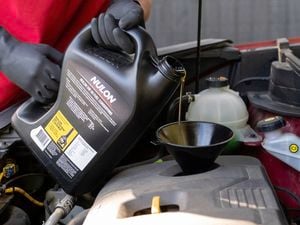 The image shows a man in a red shirt and black gloves carefully adding engine oil into a car engine using a funnel.