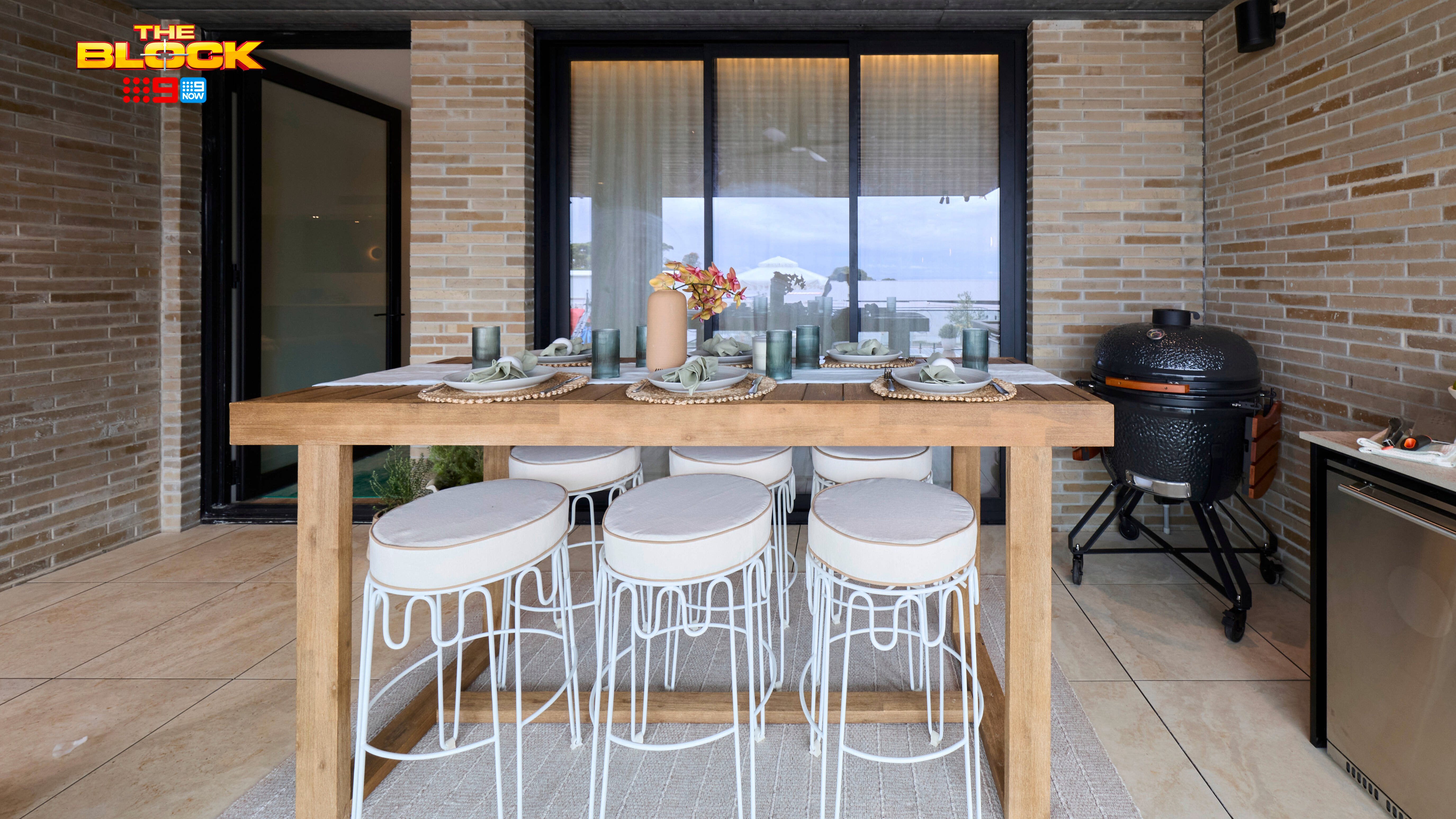 Covered outdoor dining area with a timber table, white stools, built-in barbecue and large glass doors.