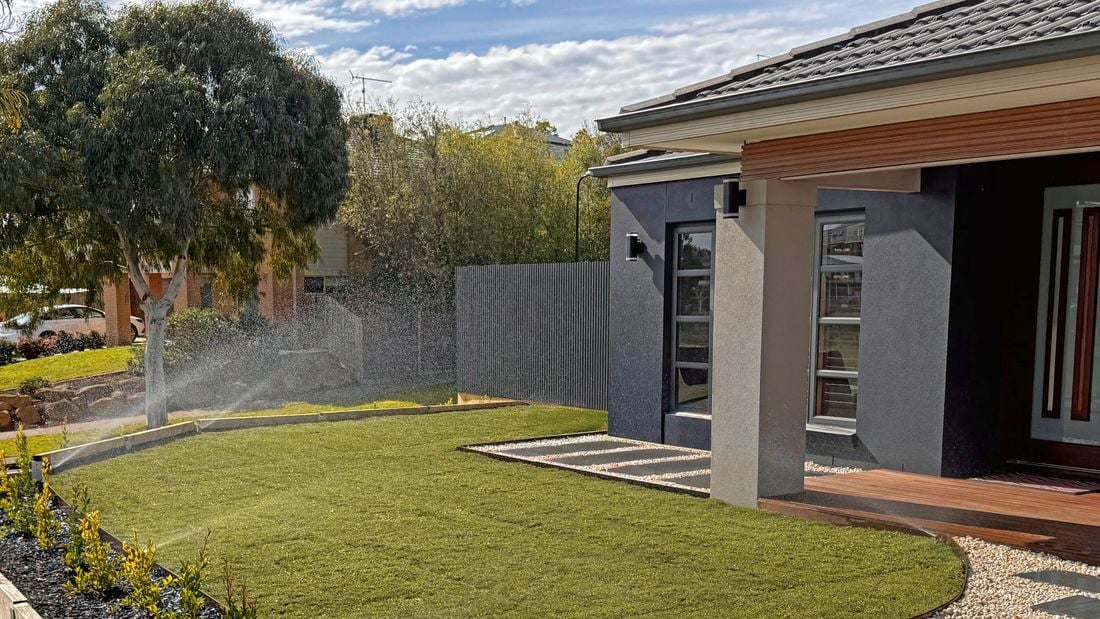 A front yard garden design with a smooth lawn being watered by pop up sprinklers in front of a modern grey house with a timber patio, and a pebbled and paved path.