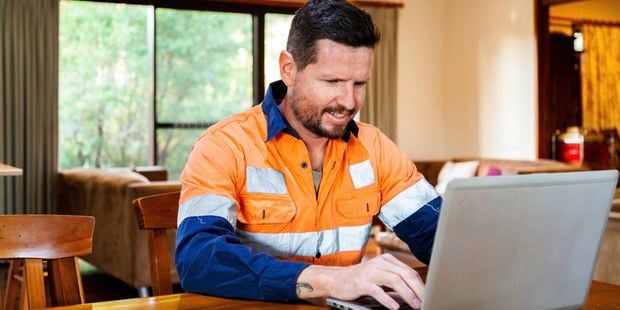 A man in hi-vis working on a laptop placed on a dining table inside a home