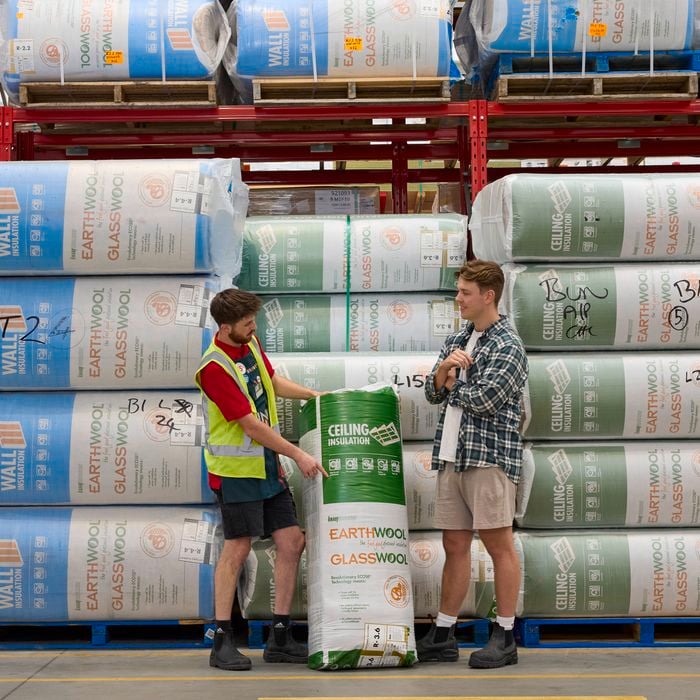 Two people review a green roll of ceiling insulation in a Bunnings warehouse aisle with stacked wall and glasswool insulation packs on shelving behind them.
