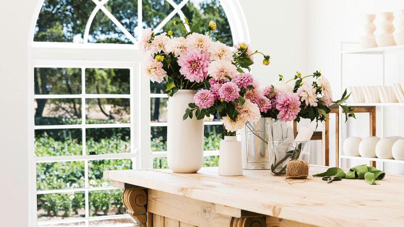 A light wooden workbench with decorative corbels, topped with vases of pink and white flowers, twine and scissors, set against a large arched window