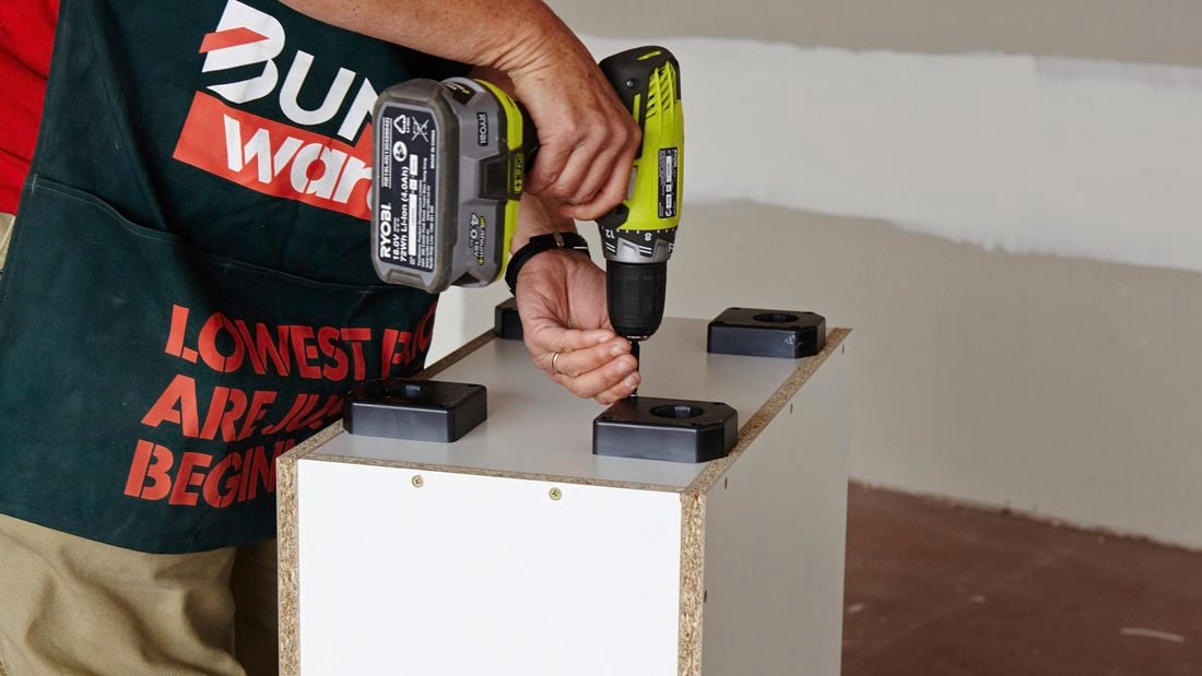 A person attaching a base plate for an adjustable foot underneath a cabinet