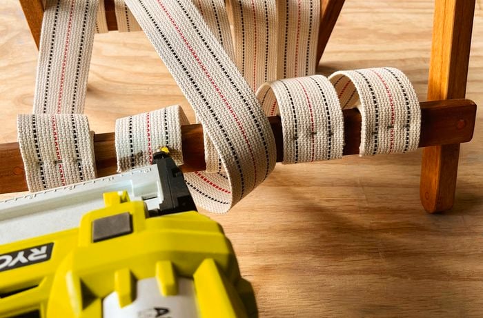 Strips of webbing looped through a wooden chair frame being tacked onto the frame with staples.