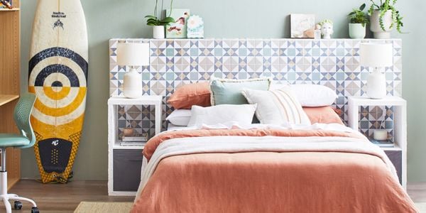  A light-coloured tiled D.I.Y. bedhead with green plants on top, behind a bed in a room with a surfboard, chair and rug