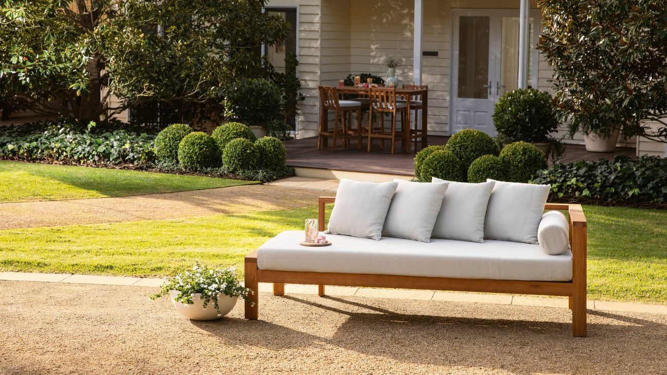   Wooden outdoor daybed with light gray cushions and striped pillows placed on a gravel path, with a small white planter beside it, set in a landscaped garden featuring manicured hedges and trees, and a house with a covered porch and dining table in the background.