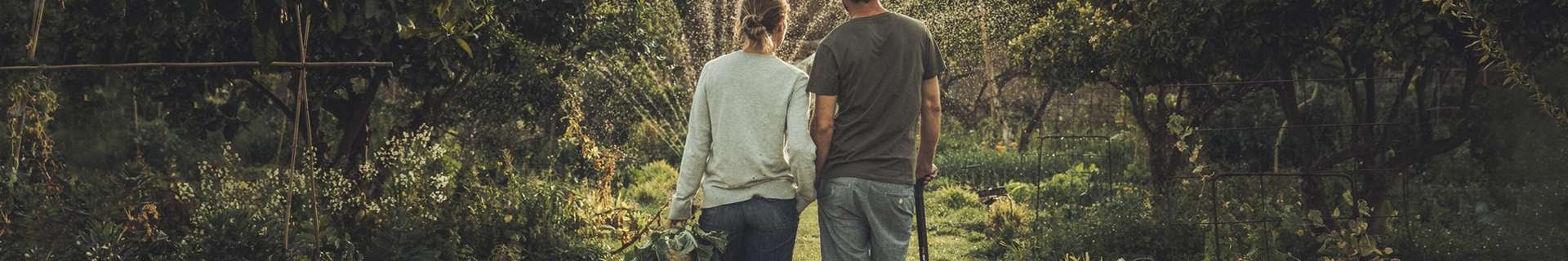 Couple walking in a garden with the sprinklers on. the man on the right is holding an axe and the woman on the left is holding picked produce