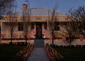 Cream traditional Australian style home with warm fairy lights across the roof, veranda and bushes along with light up candy canes as well as red and white decorative lollipops.