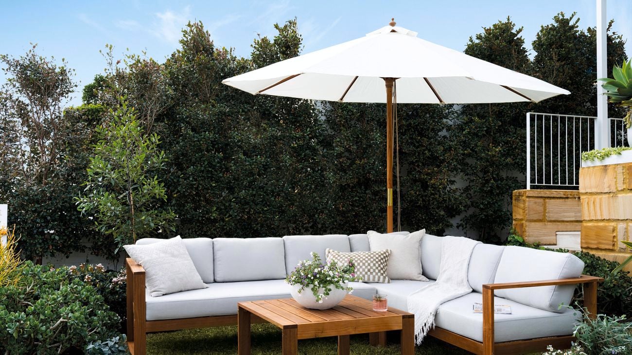 An L-shaped white outdoor lounge set with wooden frame and coffee table in a garden, with a shade umbrella propped behind.