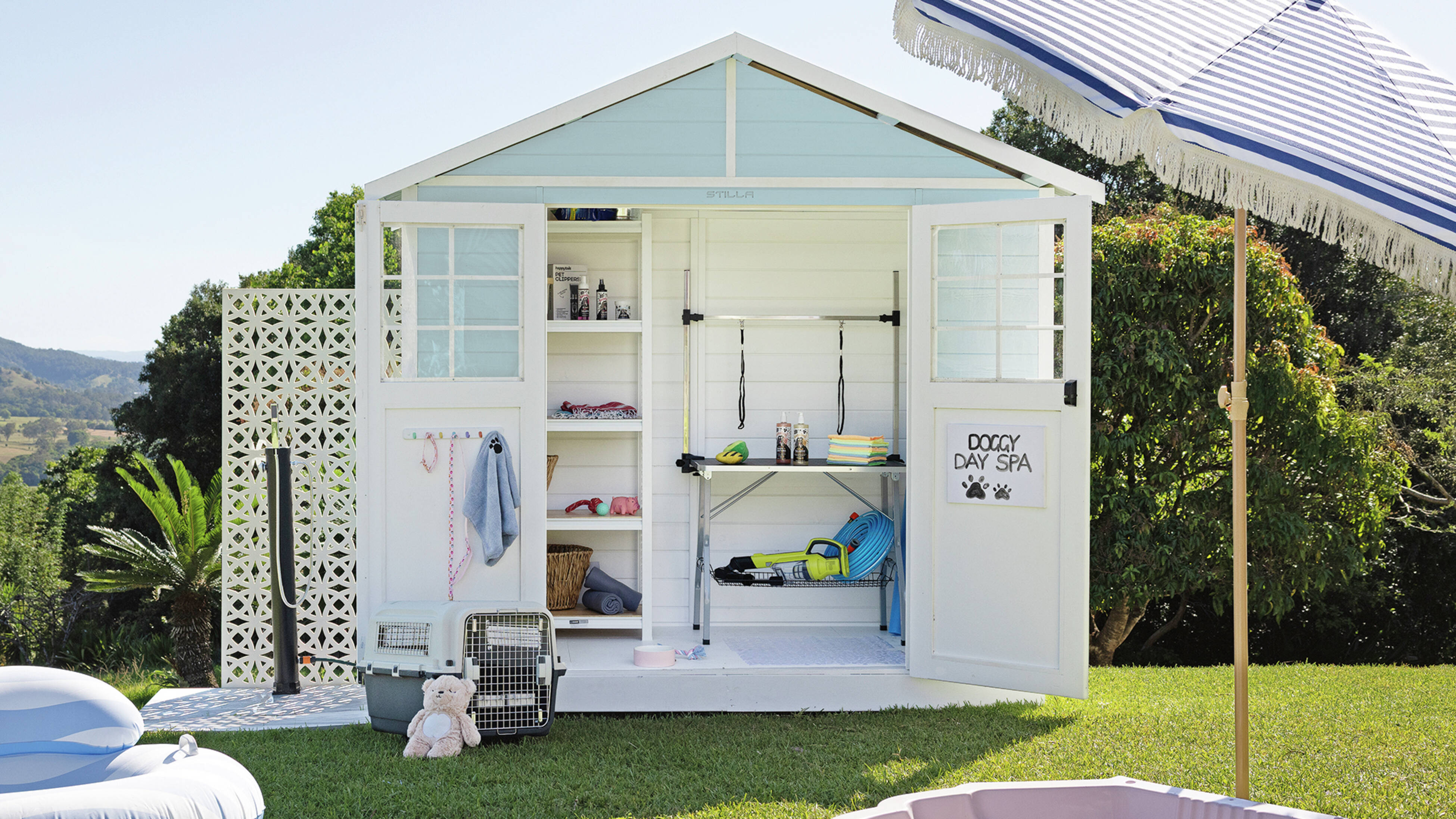 Light‑blue and white backyard shed with open doors and a sign “DOGGY DAY SPA” inside are shelves, a grooming table with straps, and pet supplies. On the grass are a pink shell paddling pool, pet carriers, a teddy bear and umbrella.