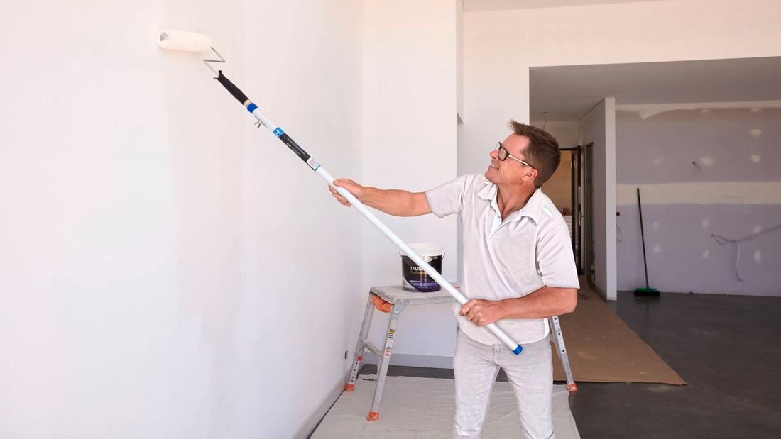 A painter dressed in white clothes uses a roller to paint a white room