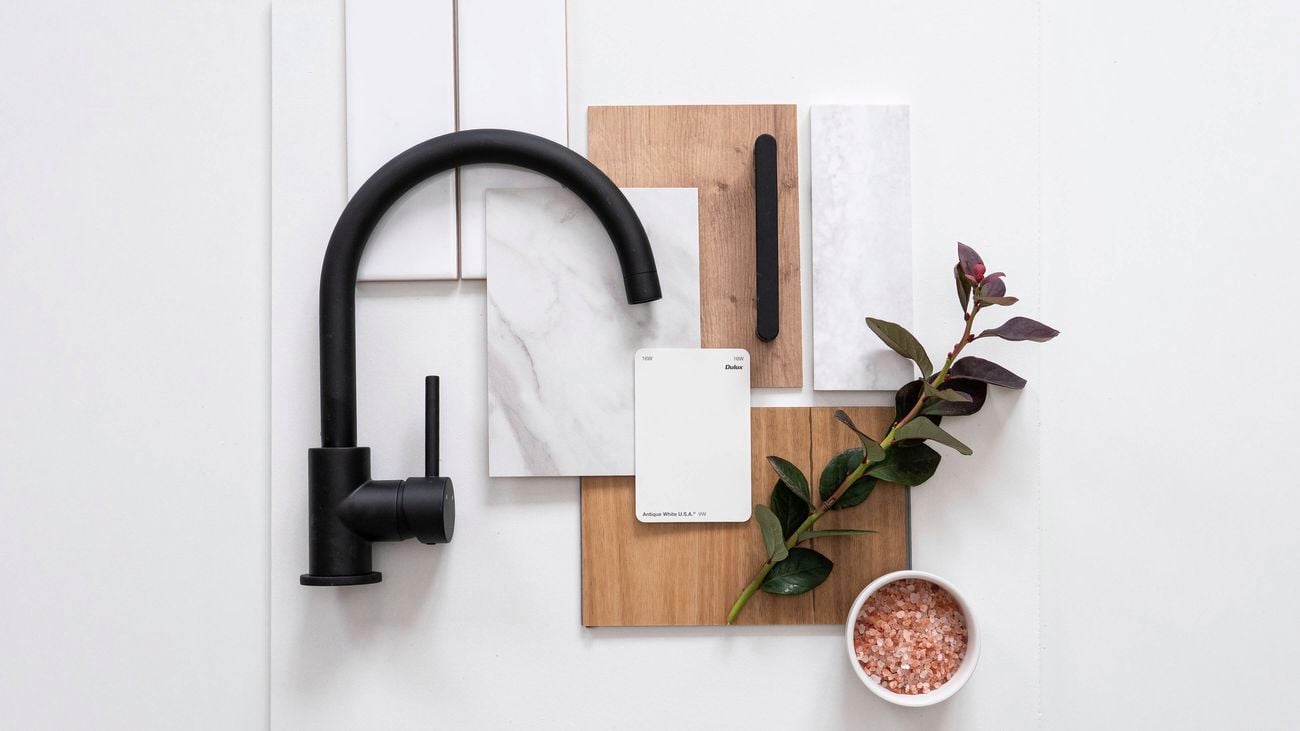 A kitchen flatlay featuring a matte black tap, white and marble tiles, light oak samples, antique white paint swatch, greenery, and a bowl of pink salt.