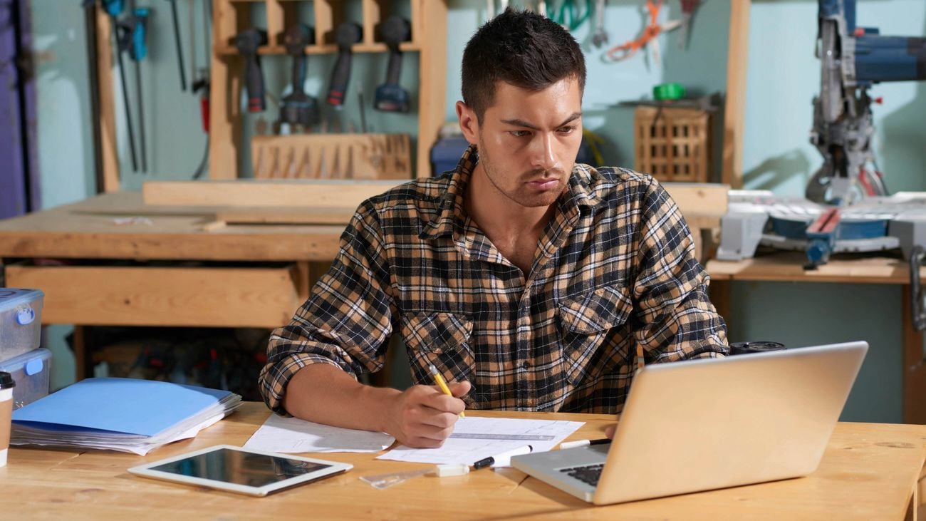 A man in a plaid shirt works on a laptop and draws on paper at a desk in a workshop.