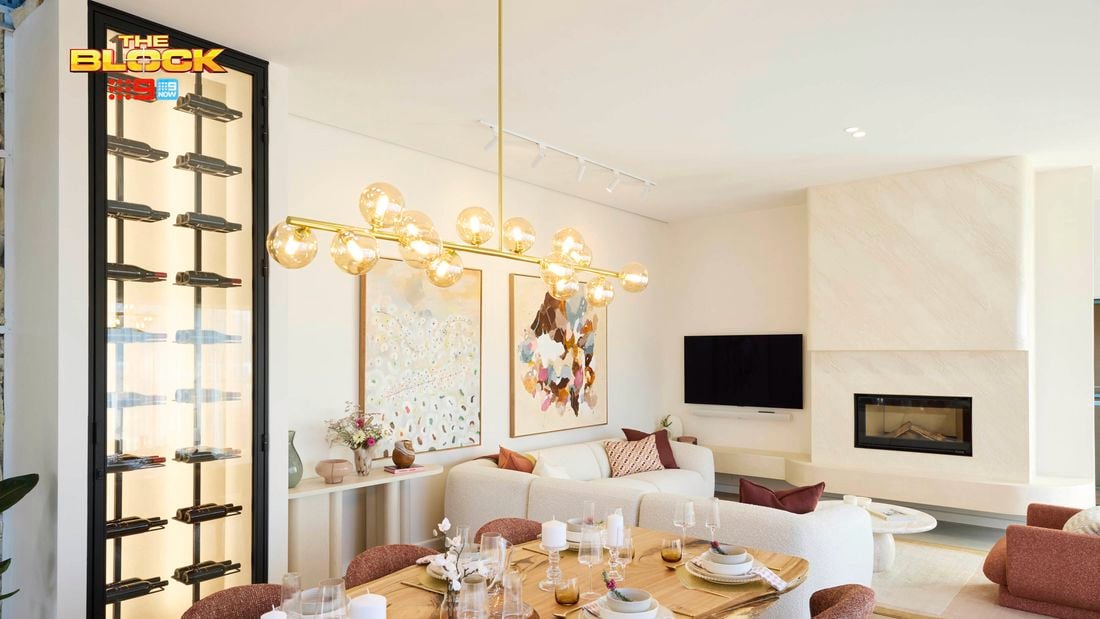 A modern living and dining area featuring a dining table set with plates, glasses, and candles, surrounded by six chairs. A contemporary chandelier with spherical lights hangs above. In the background, a white sofa with colorful cushions sits beneath two large abstract artworks. A fireplace with a TV above it is on the right, and a tall wine rack filled with bottles is on the left. 