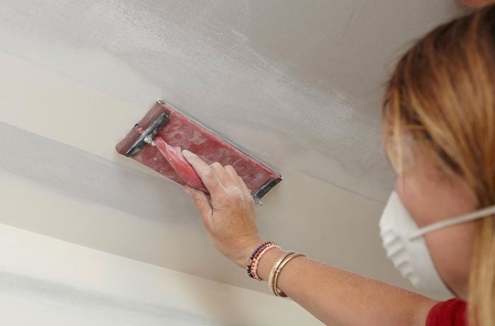 A person wearing a mask sanding a join in a plaster ceiling