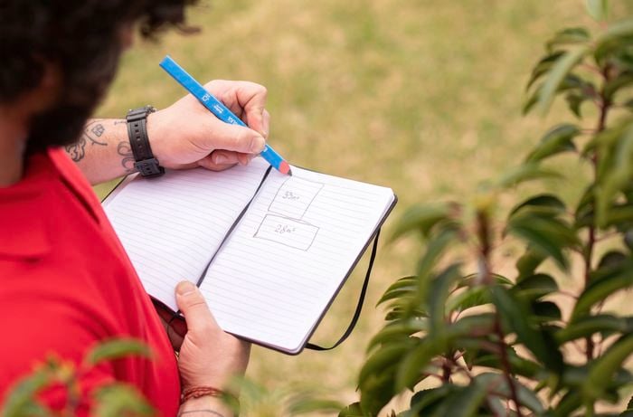 A person in a red shirt is drawing labeled diagrams calculated square meters areas for each section in a notebook while standing outdoors.