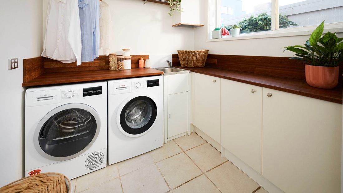 Laundry room with front-loading washer and dryer under a wooden benchtop, hanging shirts on a rail above, white cupboards, a sink in the corner, and a large window letting in natural light with indoor plants.