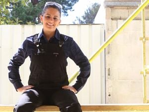 A female tradesperson wearing black Green Hip overalls sits on scaffolding on a job site.