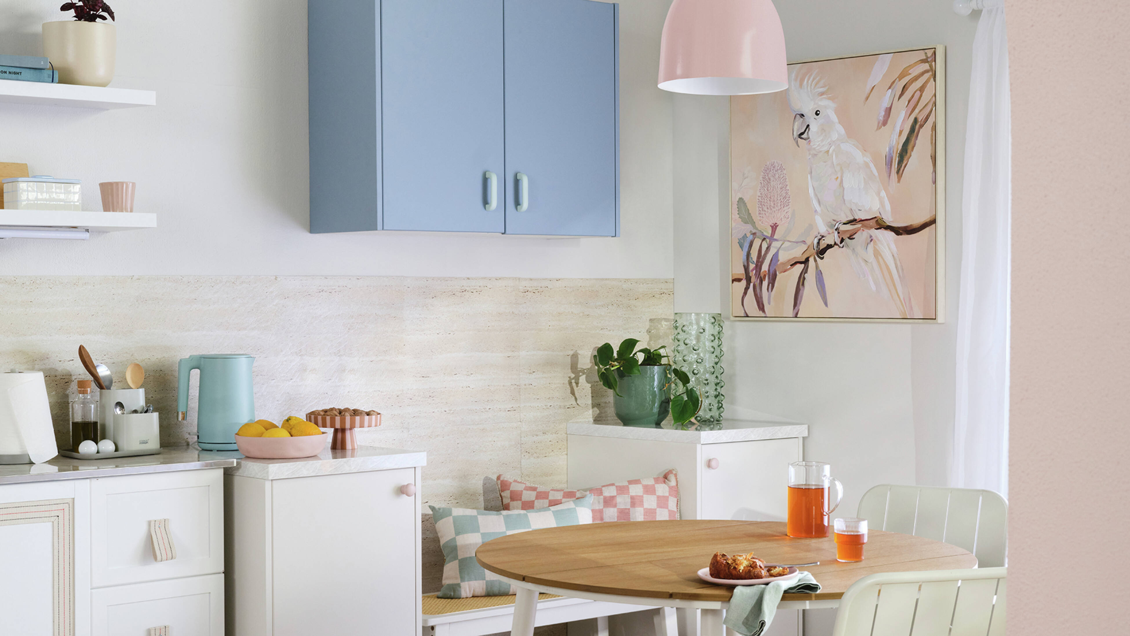 The corner of a kitchen, decked out in pastel colour tones – including a pale blue wall cabinet – with a small dining table and a cockatoo painting on the wall.