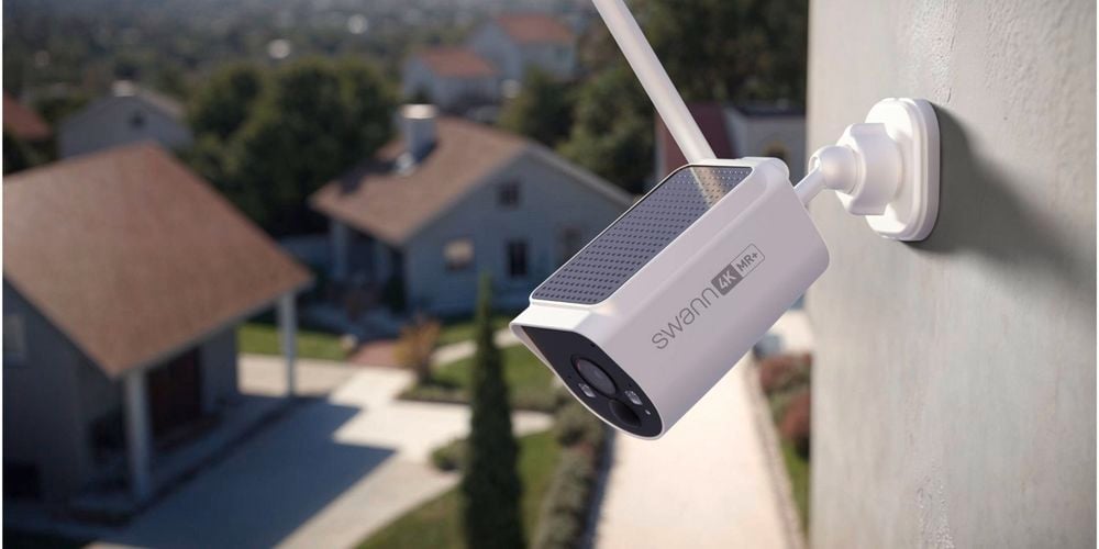 A security camera mounted on the side of a building, overlooking suburban houses and a tree-lined street in the background.