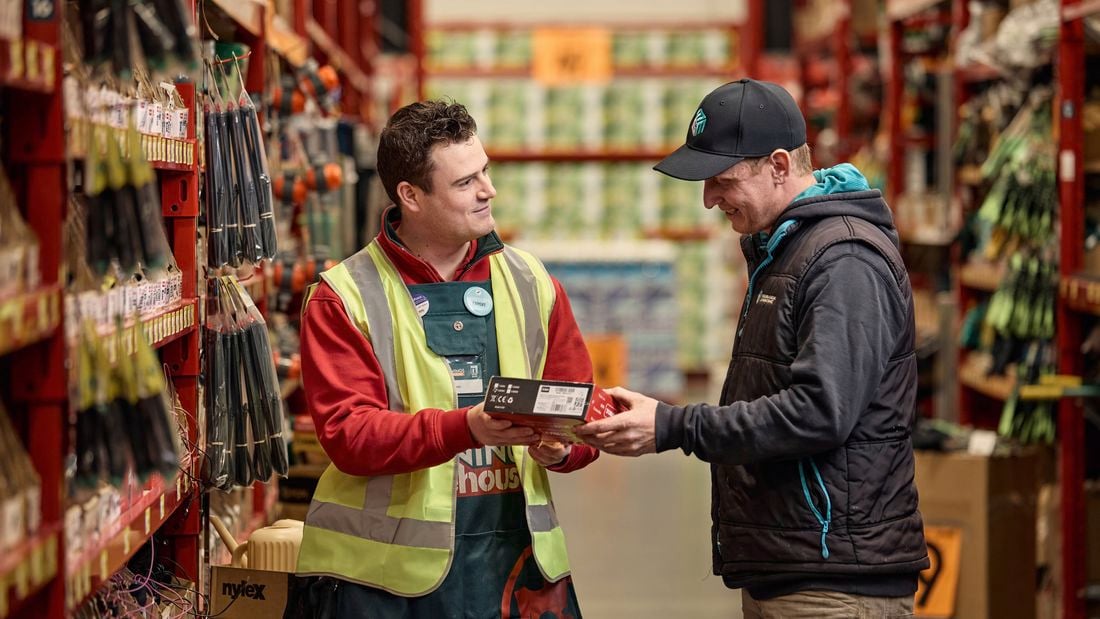 Bunnings team member handing a boxed product to a customer in a hardware store aisle with tools displayed on shelving.