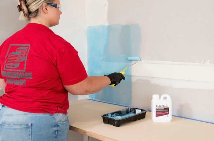 A Bunnings Team Member wearing gloves and safety glasses is using a paint roller to apply tile primer to a wall.