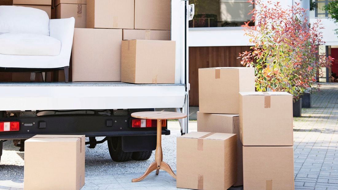  An open moving truck outside a modern house, filled with cardboard boxes and a white couch. Boxes and a small wooden table sit on the driveway.