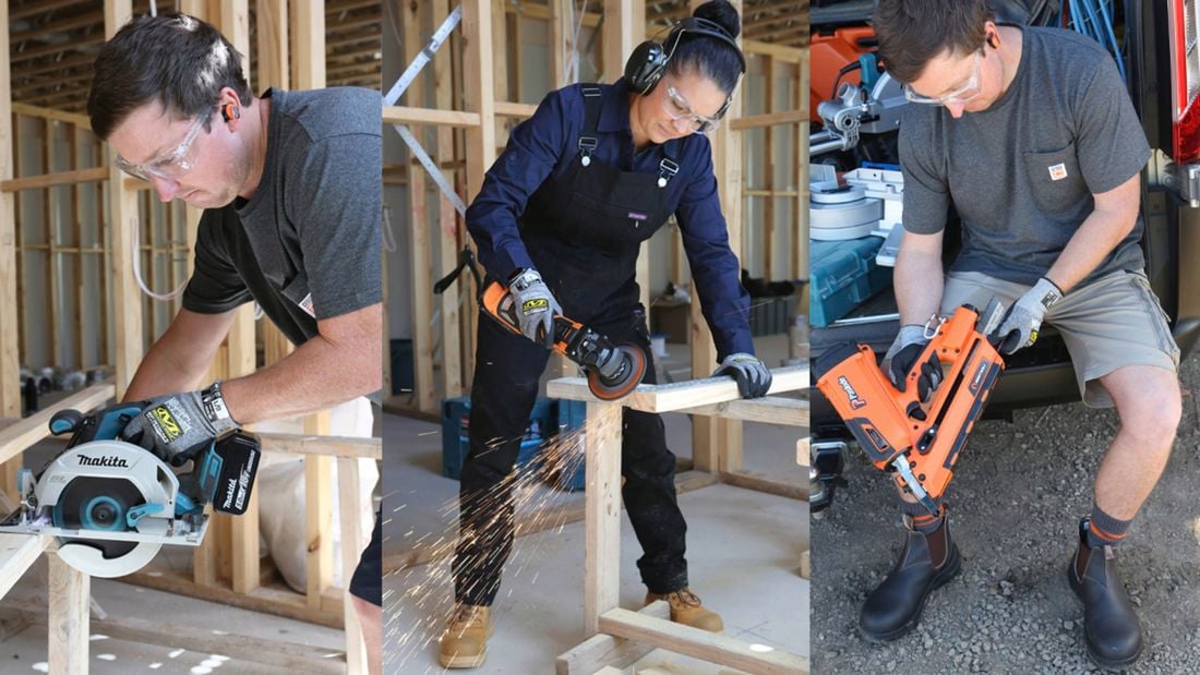 Three workers wearing safety glasses and hearing protection use power tools to cut and shape wood inside a timber-framed building.