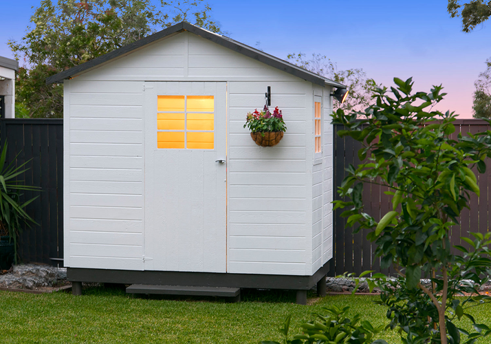 Garden shed painted white with gable roof and windows illuminated from inside