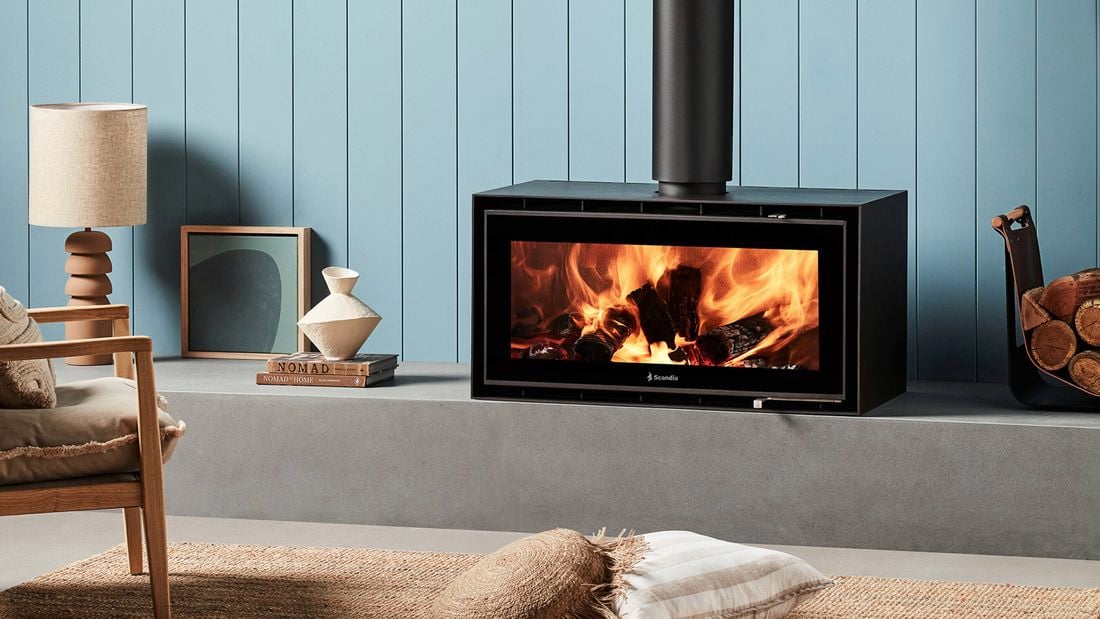 Black freestanding wood heater with burning logs against a light blue wall, next to a log holder with a rug and cushion on the floor.