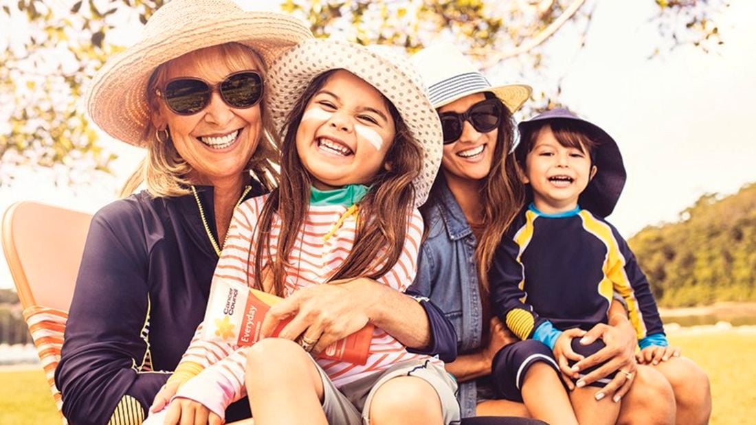 Two women both with young children, all wearing sun-smart clothing and hats, sitting outdoors. Adults wear sunglasses and children have zinc cream on their faces. One woman holds a tube of sunscreen.