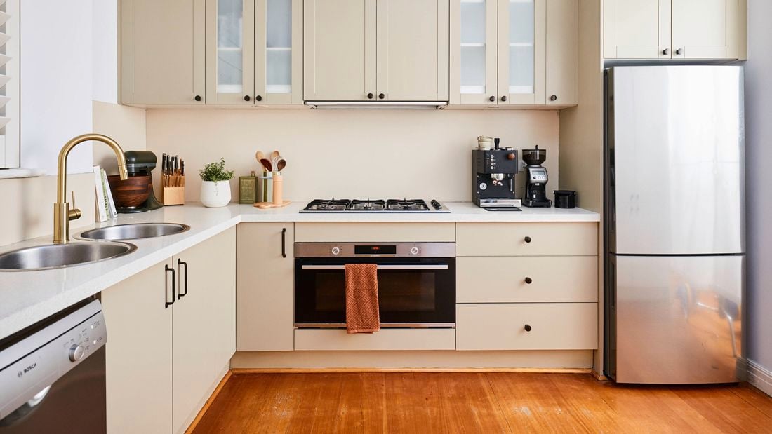 Modern U-shaped kitchen with beige cabinetry, white benchtops, and a built-in oven beneath a gas cooktop, featuring a double sink with a brass tap, small appliances on the counter, and a stainless steel refrigerator.