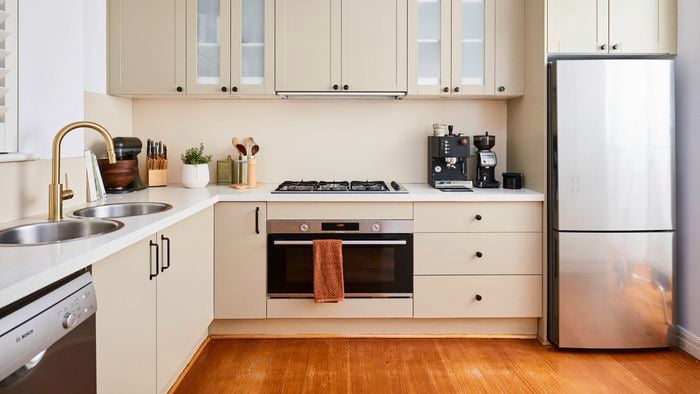 Modern U-shaped kitchen with beige cabinetry, white benchtops, and a built-in oven beneath a gas cooktop, featuring a double sink with a brass tap, small appliances on the counter, and a stainless steel refrigerator.
