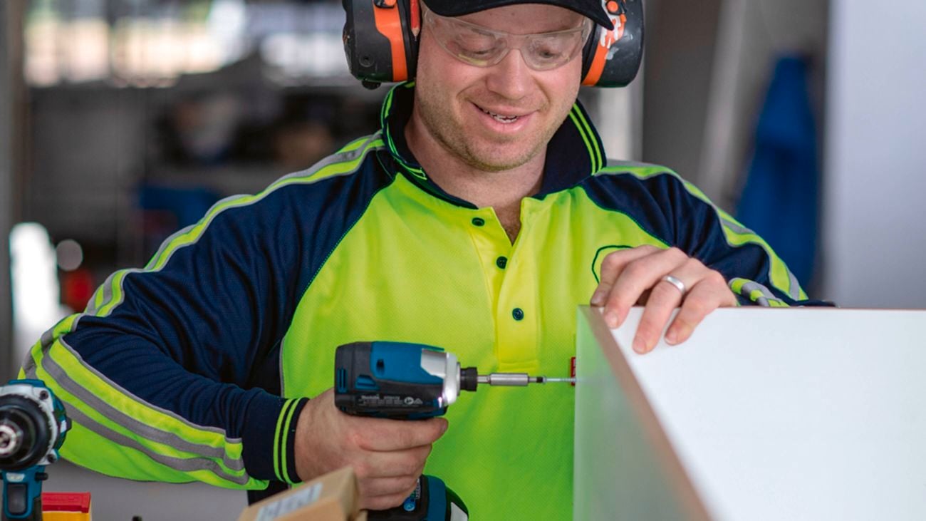 A close-up image of a man wearing a hi-vis long-sleeved shirt, safety glasses and ear protection drills into a piece of wood using a Makita hammer drill