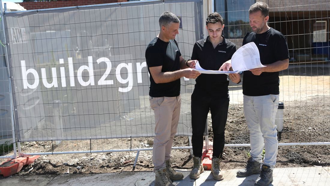 Three workers in work boots and dark shirts stand on a footpath next to a temporary construction fence, holding a large paper plan together. Muddy ground and a partially built brick house are behind the fence.]