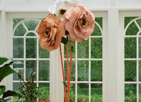 Three giant paper flowers in peach and white colours, surrounded by potted plants in a sunlit room with large windows.