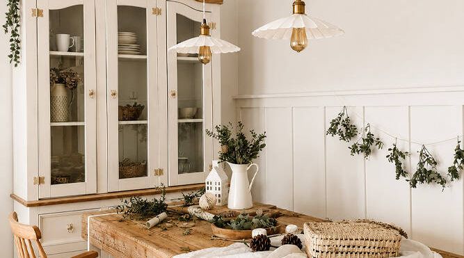 Farmhouse-style display cabinet and dining table set with white and timber finishes, pendant lighting and decorative greenery.